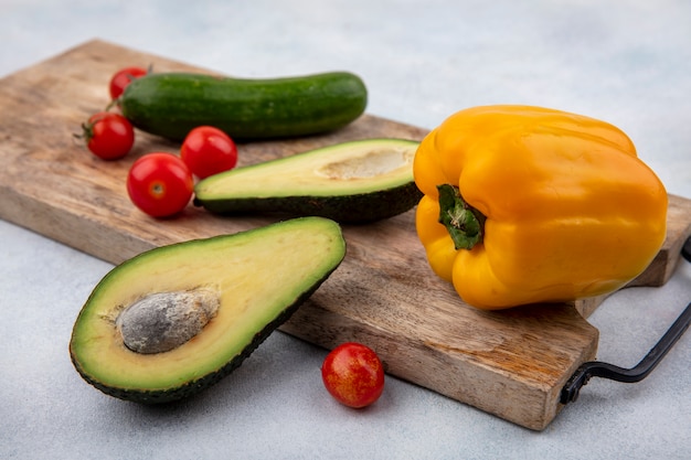 Seitenansicht der frischen Avocado-Scheiben auf einem hölzernen Küchenbrett mit Kirschtomatengurke und gelbem Paprika auf weißer Oberfläche