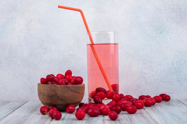 Seitenansicht der frischen roten Kornelkirschenbeeren auf einer hölzernen Schüssel mit Kornelkirschenbeersaft auf einem Glas mit Kornelkirschenbeeren lokalisiert auf einem grauen Hintergrund