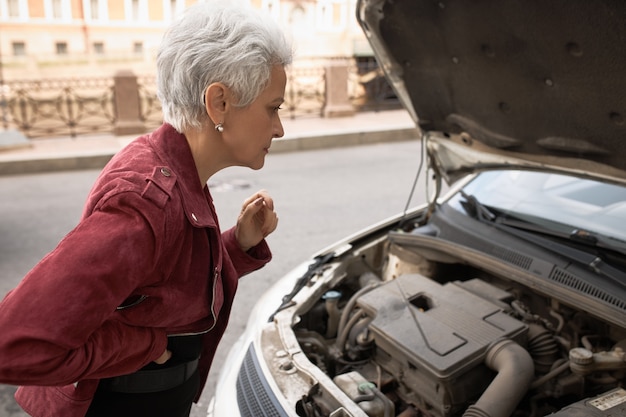 Seitenansicht der frustrierten frau mittleren alters, die mit offener motorhaube an ihrem auto steht, nach innen schaut und versucht, herauszufinden, was das problem ist.