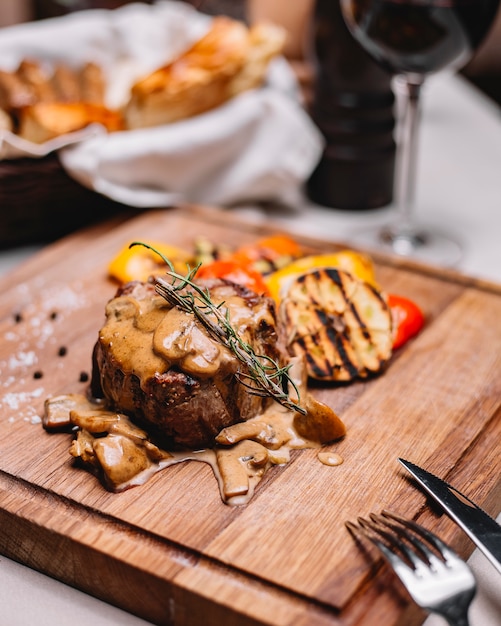 Seitenansicht der gegrillten Fleischmedaille mit Soße und gegrilltem Gemüse auf einem Holzbrett