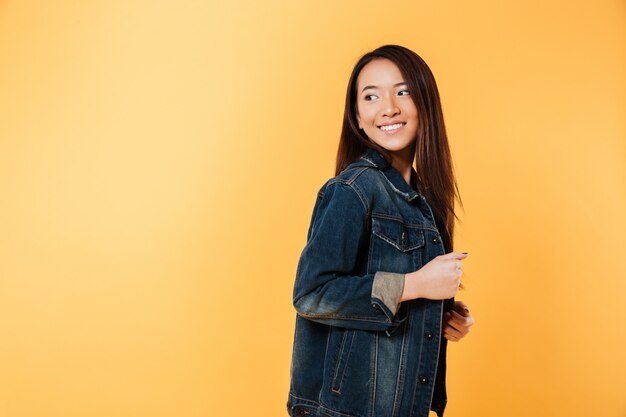 Seitenansicht der glücklichen asiatischen Frau in der Denimjacke, die zurück über gelbem Hintergrund aufwirft und schaut