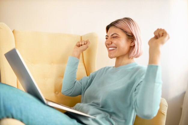 Seitenansicht der glücklichen emotionalen Freiberuflerin der jungen Frau in der Freizeitkleidung, die im Sessel mit tragbarem Computer auf ihrem Schoß sitzt, Fäuste ballt, über großes Arbeitsangebot aufgeregt ist, ausrufend