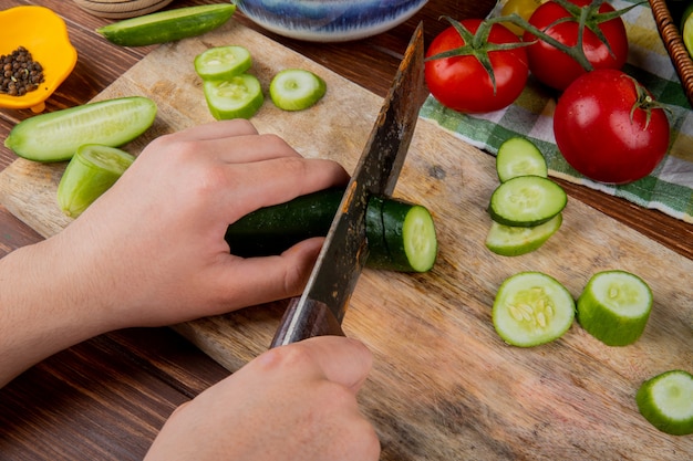 Seitenansicht der Hände, die Gurke mit Messer auf Schneidebrett mit Tomaten schwarzem Pfeffer auf Holzoberfläche schneiden