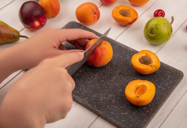 Seitenansicht der Hände, die Pfirsich mit Messer und halb geschnittenem Pfirsich auf Schneidebrett mit Muster von Birnenpfirsichen auf hölzernem Hintergrund schneiden