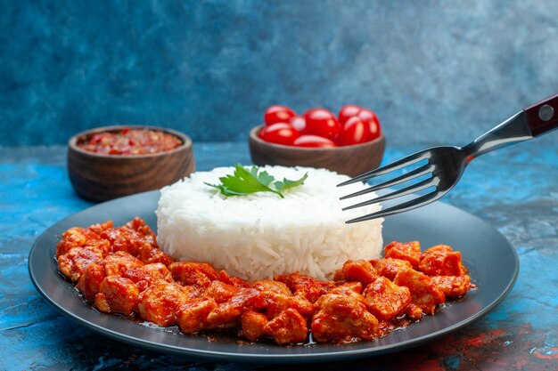 Seitenansicht der Hand, die eine Gabel auf Reismehl mit Hühnchen und Sauce auf einem schwarzen Teller und Tomaten auf blauem Hintergrund hält