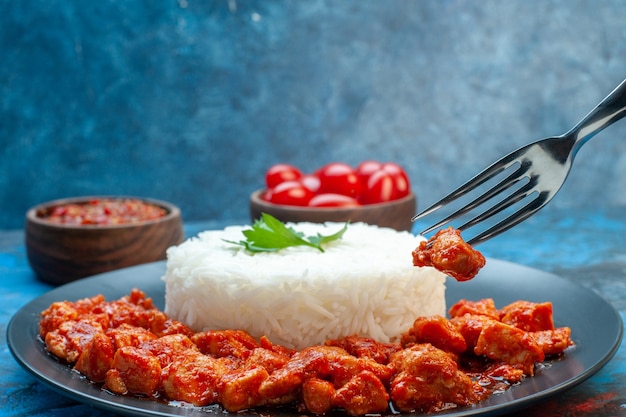 Seitenansicht der Hand, die eine Gabel hält, die Fleisch aus Reismehl mit Hühnchen und Sauce auf einem schwarzen Teller und Tomaten auf blauem Hintergrund nimmt