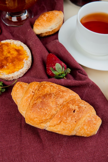 Seitenansicht der Hörnchenrolle mit knuspriger Knäckebroterdbeere auf Bordotuch mit Tasse Tee auf weißem Tisch