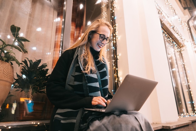 Seitenansicht der jungen Frau, die Laptop auf Straße mit Nachtstadt verwendet