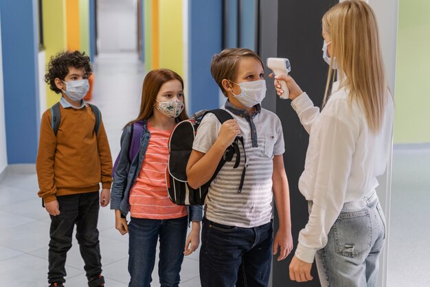 Seitenansicht der Lehrerin mit medizinischer Maske, die die Temperatur der Kinder in der Schule prüft