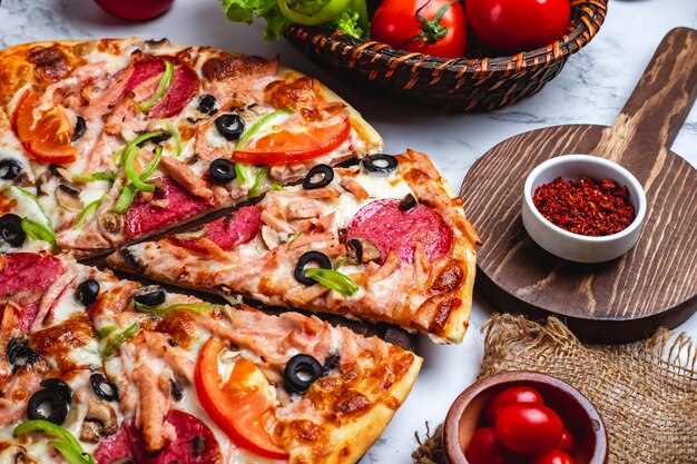 Seitenansicht der Pizza mit Salami-Schinken-Paprika-Tomaten, schwarzen Oliven und Käse auf dem Tisch