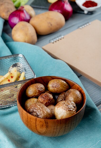 Seitenansicht der Schüssel der gebackenen Kartoffeln auf Stoff mit Zwiebeln und Kartoffeln auf hölzernem Hintergrund mit Kopienraum