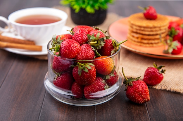 Seitenansicht der Schüssel Erdbeere mit Tasse Tee und Zimt- und Waffelkeksen auf Sackleinen und Holzoberfläche