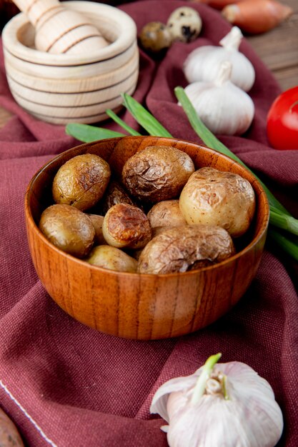 Seitenansicht der Schüssel voll Ofenkartoffel mit Gemüse und Knoblauchbrecher herum auf burgunderfarbener Stoffoberfläche