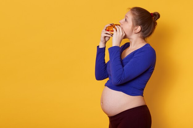 Seitenansicht der schwangeren Dame isst Fast Food, trägt blaues Hemd und Leggins, mit Bündel auf dem Kopf