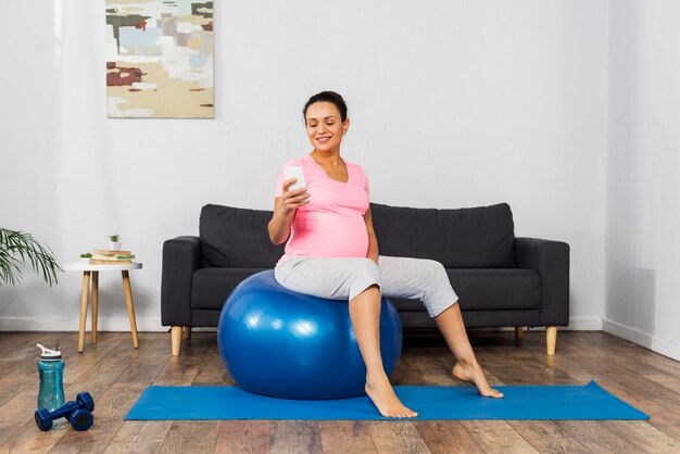 Seitenansicht der schwangeren Frau des Smileys zu Hause unter Verwendung des Smartphones und des Trainings mit Ball