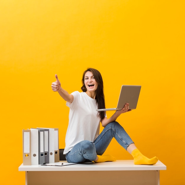 Kostenloses Foto seitenansicht der smileyfrau sitzend auf schreibtisch und daumen beim halten des laptops aufgebend