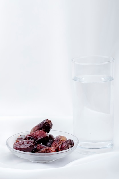 Seitenansicht der süßen getrockneten dattelfrüchte in einer untertasse mit einem glas wasser auf weißem hintergrund