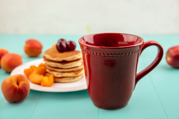 Seitenansicht der Tasse Kaffee mit Pfannkuchenplatte und Aprikosenscheiben mit Kirschen auf blauer Oberfläche und weißem Hintergrund