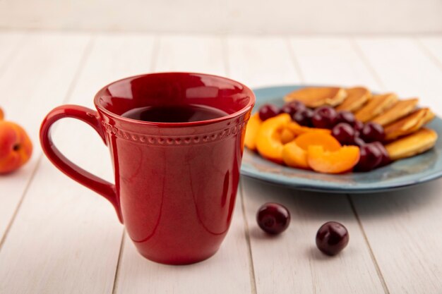 Seitenansicht der Tasse Kaffee mit Teller Pfannkuchen und Aprikosenscheiben mit Kirschen auf hölzernem Hintergrund