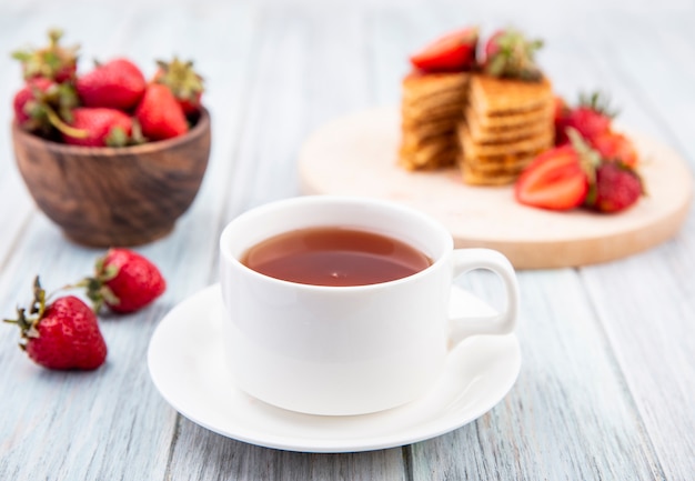Seitenansicht der Tasse Tee auf Untertasse und Waffelkekse mit Erdbeeren in Teller und Schüssel auf Holzoberfläche