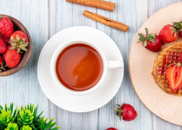 Seitenansicht der Tasse Tee auf Untertasse und Waffelkekse mit Erdbeeren in Teller und Schüssel mit Zimt auf Holzoberfläche