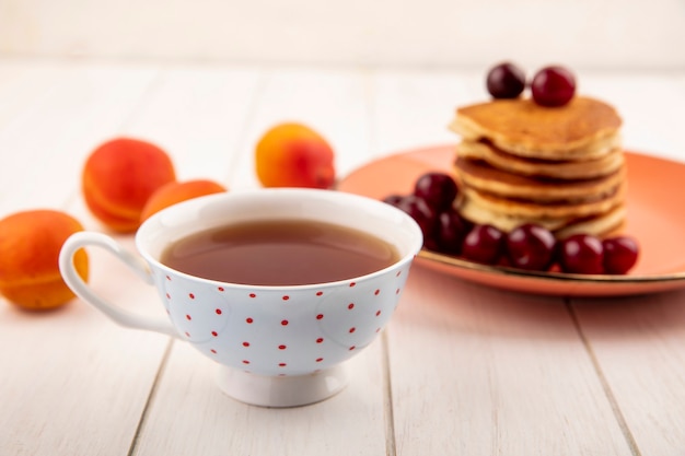 Seitenansicht der Tasse Tee mit Teller mit Pfannkuchen und Kirschen und Aprikosen auf hölzernem Hintergrund