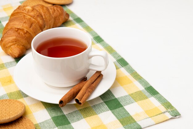 Seitenansicht der Tasse Tee mit Zimt auf Teebeutel und Kekse mit japanischer Butterrolle auf kariertem Stoff auf weißem Hintergrund mit Kopienraum