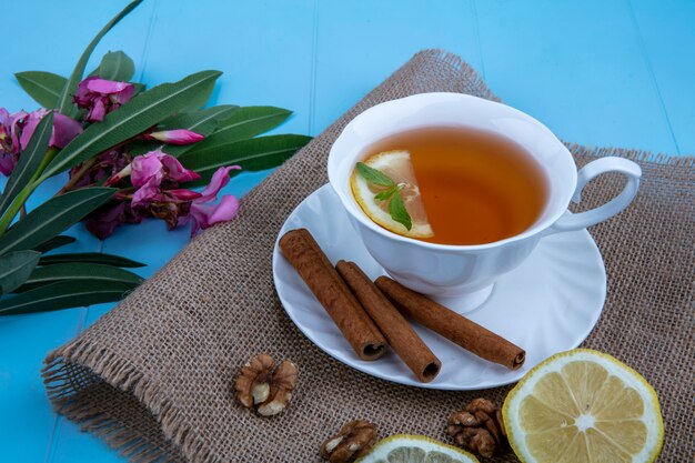 Seitenansicht der Tasse Tee mit Zitronenscheibe und Zimt auf Untertasse mit Walnuss-Zitronenscheiben auf Sackleinen mit Blumen und Blättern auf blauem Hintergrund