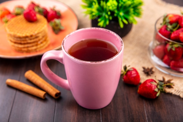Seitenansicht der Tasse Tee und Waffelkekse mit Erdbeeren in Teller und Schüssel auf Sackleinen mit Zimt auf Holzoberfläche