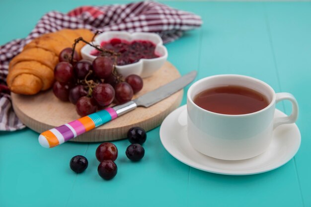 Seitenansicht der Traube des Croissants und der Himbeermarmelade mit Messer auf Schneidebrett auf kariertem Stoff und Tasse Tee auf blauem Hintergrund