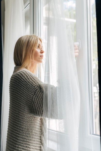 Seitenansicht der traurigen Frau zu Hause während der Pandemie, die durch das Fenster schaut