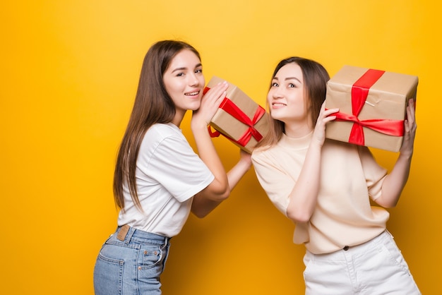 Seitenansicht der überraschten jungen Frauen halten Geschenkbox mit Geschenkbandschleife lokalisiert auf gelber Wand. Frauentag Geburtstag, Feiertagskonzept.