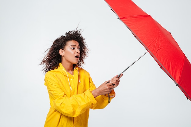 Seitenansicht der verwirrten afrikanischen Frau im Regenmantel, der Regenschirm hält