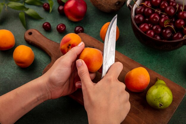 Seitenansicht der weiblichen Hände, die Aprikose mit Messer und Birne auf Schneidebrett und Kirschen in Schüssel mit Pfirsich-Kokosnuss und Blättern auf grünem Hintergrund schneiden