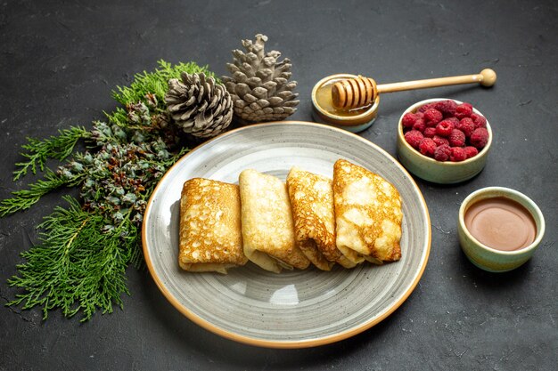 Seitenansicht des Abendessenhintergrundes mit köstlichem Pfannkuchenhonig und Schokoladenhimbeer- und Nadelbaumkegel auf schwarzem Hintergrund