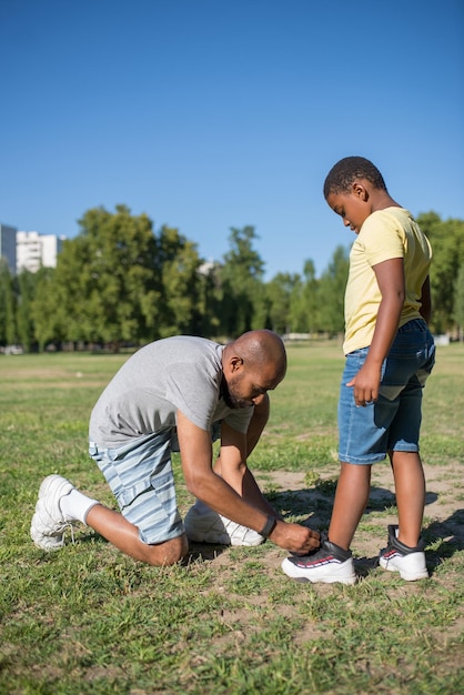 Seitenansicht des afroamerikanischen Mannes und des süßen Jungen, der auf dem Feld steht. Ernster Vater, der auf seinem einen Knie auf Gras steht und auf Kinderturnschuh schaut, der Schnürsenkel bindet. Elternbetreuung und sportliches Aktivitätskonzept