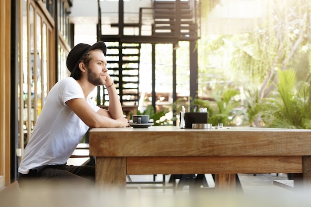 Seitenansicht des attraktiven jungen Hipsters im Hut, der allein in der Straßencafeteria sitzt und seinen Ellbogen auf massivem Holztisch ruht