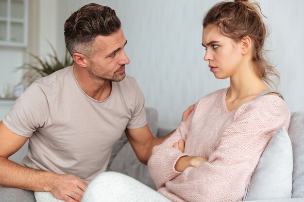 Seitenansicht des aufmerksamen Mannes sitzend auf Couch und seine ernste Freundin mit den gekreuzten Armen zu Hause beruhigend