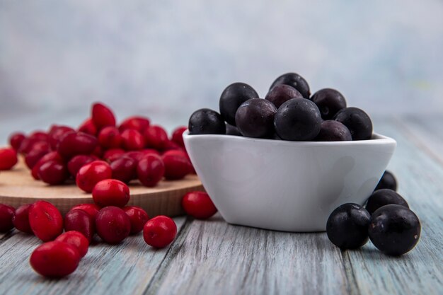 Seitenansicht des dunkelhäutigen Schwarzdorns auf einer weißen Schüssel mit roten Kornelkirschenbeeren lokalisiert auf einem hölzernen Küchenbrett auf einem grauen hölzernen Hintergrund