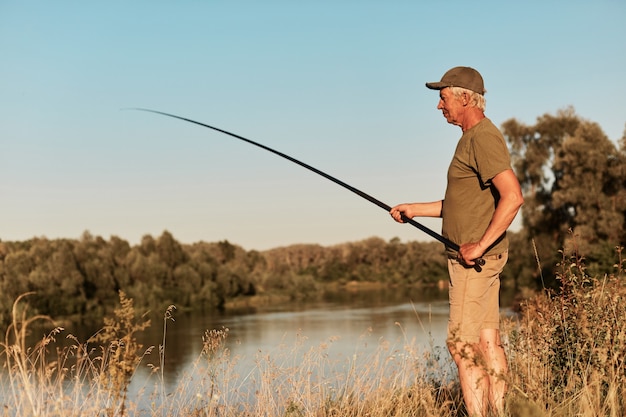 Seitenansicht des Fischers, der am Ufer des Sees oder des Flusses steht und seine Angelrute in den Händen betrachtet, auf Sonnenuntergang fischend, auf schöne Natur, grünes T-Shirt und Hose tragend.