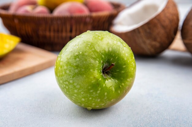 Seitenansicht des frischen und grünen Apfels mit Pfirsichen auf einem Eimer und einer halben Kokosnuss auf weißer Oberfläche