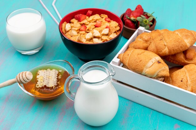 Seitenansicht des Frühstücks mit Croissants, Cornflakes, Früchten, Milch und Honig auf blauer Oberfläche horizontal