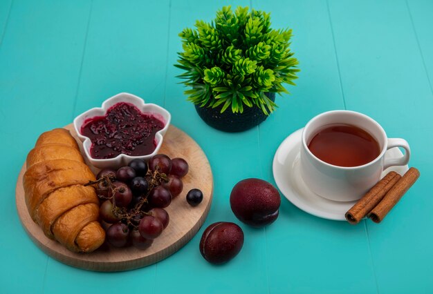 Seitenansicht des Frühstückssatzes mit Croissant- und Himbeermarmeladentraube auf Schneidebrett und Tasse Tee mit Zimt- und Pluotspflanze auf blauem Hintergrund