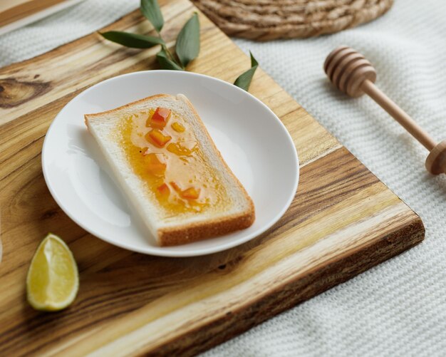 Seitenansicht des Frühstückssets mit Brotscheibe, bestrichen mit Marmelade im Teller und Limettenscheibe mit Blättern auf Schneidebrett und Marmeladenschöpflöffel auf weißem Stoffhintergrund