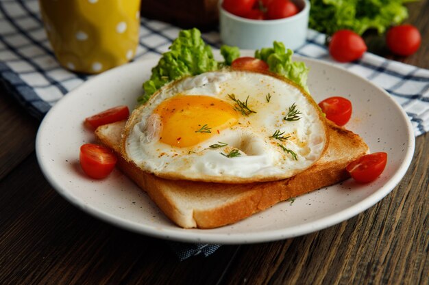 Seitenansicht des Frühstückssets mit Spiegelei-Salat-Tomaten auf getrockneter Brotscheibe im Teller mit Orangensaft auf kariertem Stoff auf Holzhintergrund