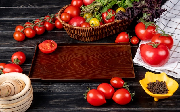 Seitenansicht des Gemüses als Tomatengrünminze-Basilikum im Korb und geschnittene Tomate im Tablett-Knoblauchbrecher des schwarzen Pfeffers auf Holztisch