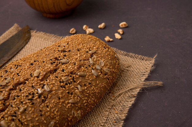 Seitenansicht des gesäten Baguettebrotes auf Sackleinen mit Körnern auf kastanienbraunem Hintergrund mit Kopienraum