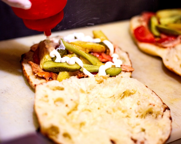 Seitenansicht des Gießens von Mayonnaise in einen Hühnerdoner in Brot mit Gurken