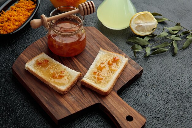 Seitenansicht des Glases mit Quittenmarmelade und mit Quittenmarmelade bestrichenen Brotscheiben auf Schneidebrett und geriebener Orangenschale Glas Limettensaft halb geschnittene Zitrone und Blätter auf schwarzem Hintergrund