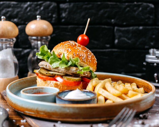 Seitenansicht des Hühnchen-Burgers mit Gurken und Tomaten, serviert mit Pommes Frites und Saucen auf Dunkelheit